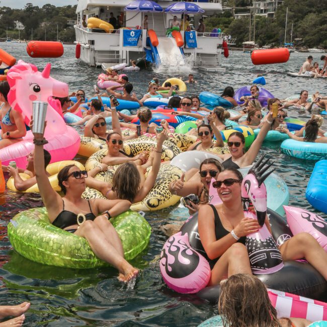 A group of people enjoys a sunny day on inflatable pool floats in the water near a party boat. They are laughing and holding drinks, surrounded by colorful floats shaped like animals and other objects, with a slide at the back of the boat.