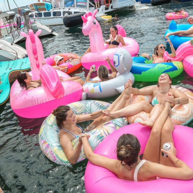 A group of people are having fun on colorful inflatable floats, shaped like unicorns and flamingos, in a marina. They're surrounded by boats, enjoying a sunny day on the water, laughing and relaxing.