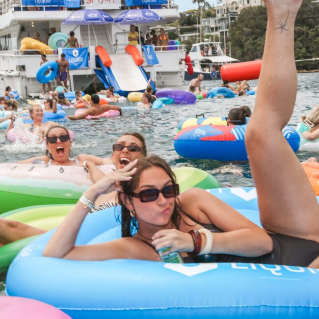 People enjoying a lively pool party on inflatable tubes in the water. A large boat with slides is in the background, and everyone seems to be having fun under a sunny sky. One person is posing playfully in the foreground.