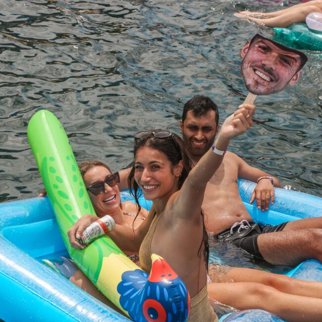 Three people are lounging on an inflatable raft in a body of water. A woman in front is holding a large cutout of a man's face. Another woman holds a drink, and a man smiles behind them. They appear to be enjoying a sunny day.