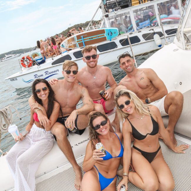 A group of six people sits and lounges on the deck of a boat, smiling and enjoying drinks. They are in swimwear, surrounded by water with other boats and people in the background on a sunny day.