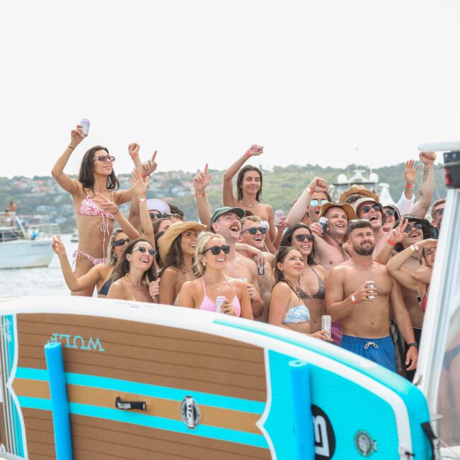 A large group of people in swimwear pose for a photo on a boat, holding drinks and cheering. A paddleboard is seen in the foreground. Other boats and a distant shoreline are visible in the background.