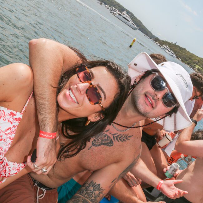 A woman in sunglasses and a floral swimsuit poses with a man wearing a white bucket hat and sunglasses. They are on a boat with others in the background. The scene is lively with water and other boats in the distance.