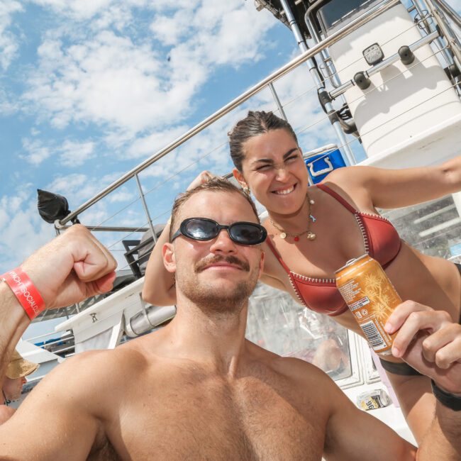 A man and a woman wearing swimsuits pose on a boat. The man raises a fist and wears sunglasses, while the woman smiles, also in sunglasses, holding a beverage can. The sky is clear with a few clouds in the background.