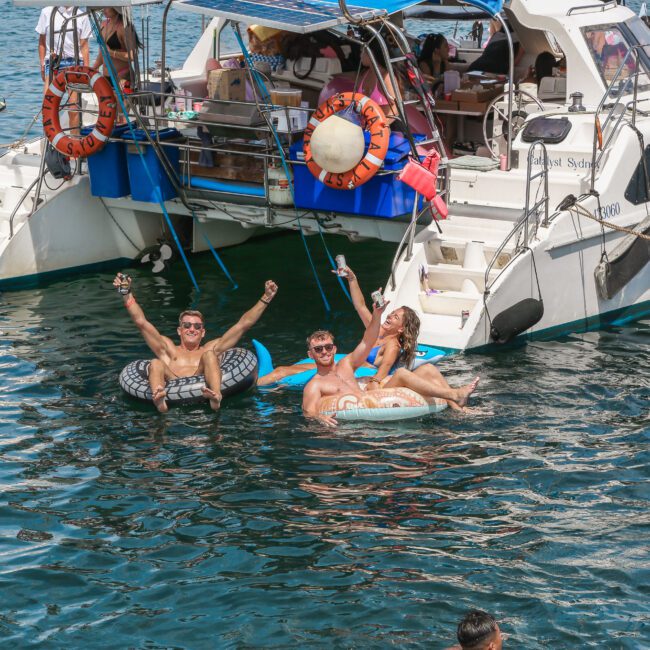 Three people float on inflatables in the water near a boat. Two hold drinks, smiling, while another lounges nearby. The boat is anchored with life rings visible, and others are on board enjoying the sunny day.