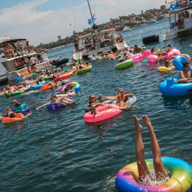 A vibrant scene on the water with people relaxing on colorful inflatable rings and floating around a few boats. One person is diving headfirst into the water from a rainbow-colored float. The sky is clear and sunny.