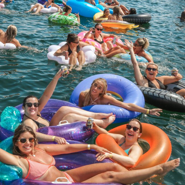 A group of people in swimsuits relax on colorful inflatable pool floats in a lake. They are smiling, holding drinks, and enjoying the sunny day. The water is calm, and more people can be seen lounging in the background.