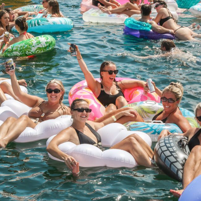 A group of people in swimsuits are floating on inflatable rafts, holding drinks, and smiling in a large body of water. The scene is lively with many colorful inflatables and others in the background enjoying the water.