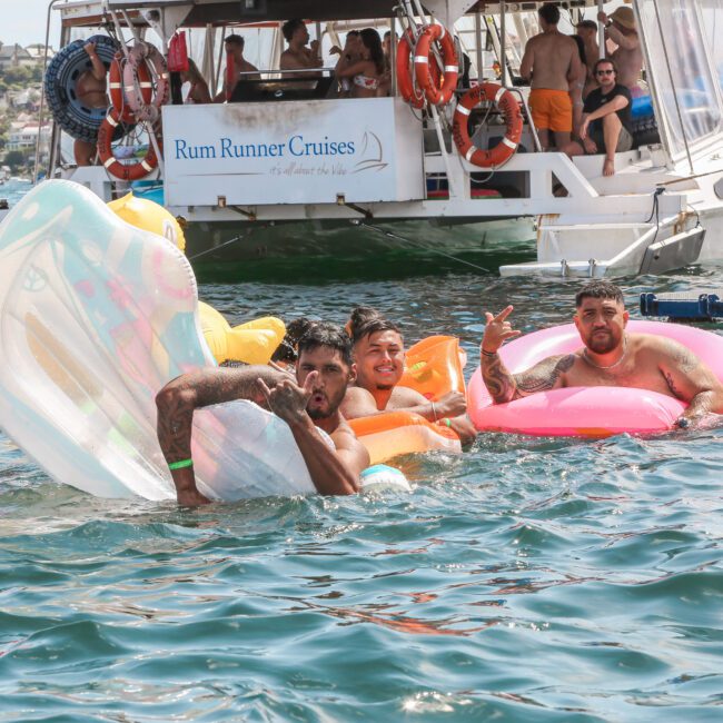 Three people in colorful inflatable rings enjoy the water near a boat labeled "Rum Runner Cruises." People are visible on the boat in the background, and the scene conveys a lively, sunny day on the water.