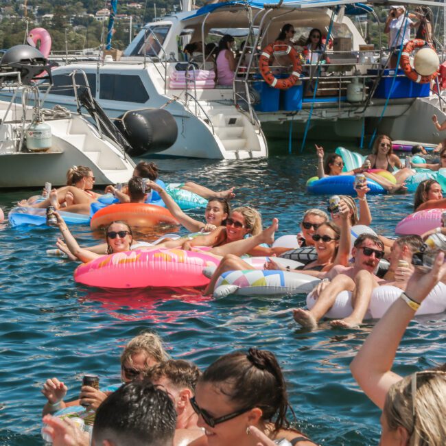 A group of people enjoying a sunny day in the water on various inflatable floats. They are smiling and holding drinks, surrounded by boats, creating a lively and festive atmosphere.