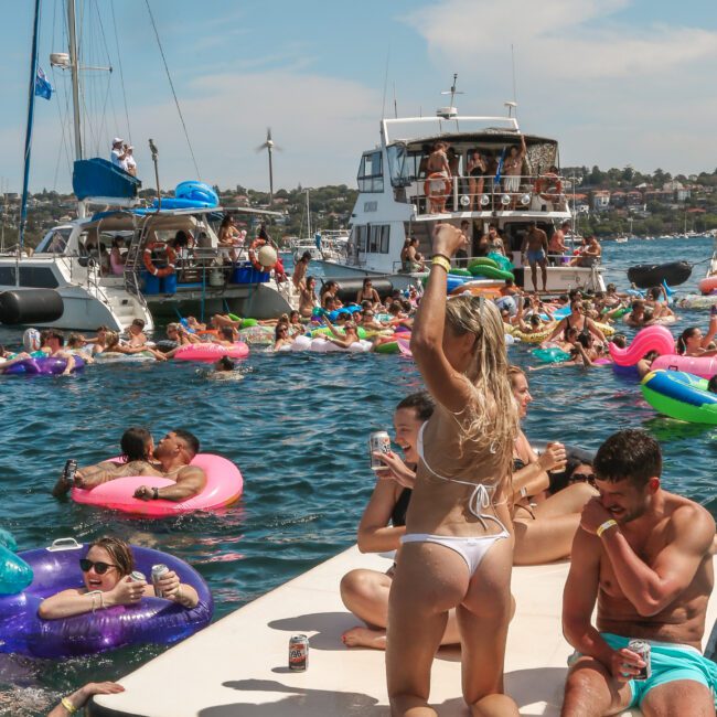 A lively scene of people enjoying a sunny day on the water, with many floating on colorful inflatables and socializing. A woman in a white bikini stands on a floating dock, holding up a drink. Several boats are in the background.