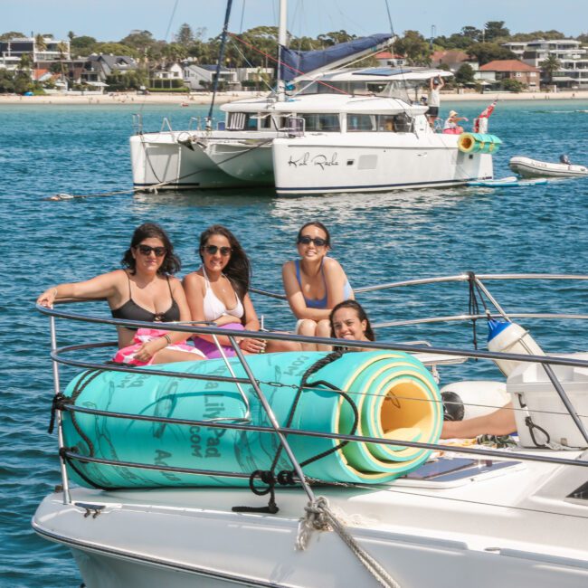 Four people relaxing on the deck of a boat, with a large rolled-up mat beside them. They are smiling and wearing sunglasses. In the background, another boat and a scenic shoreline with buildings and trees are visible. The water is calm.