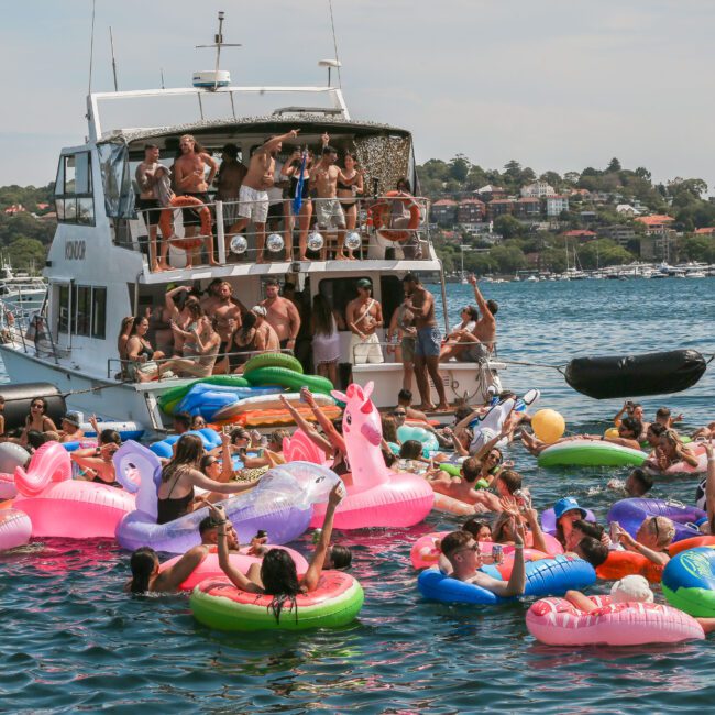 A lively scene of people enjoying a sunny day on inflatable floats in the water, near a docked boat. The boat is crowded with more people partying. The sky is clear, with a backdrop of trees and houses on the shore.