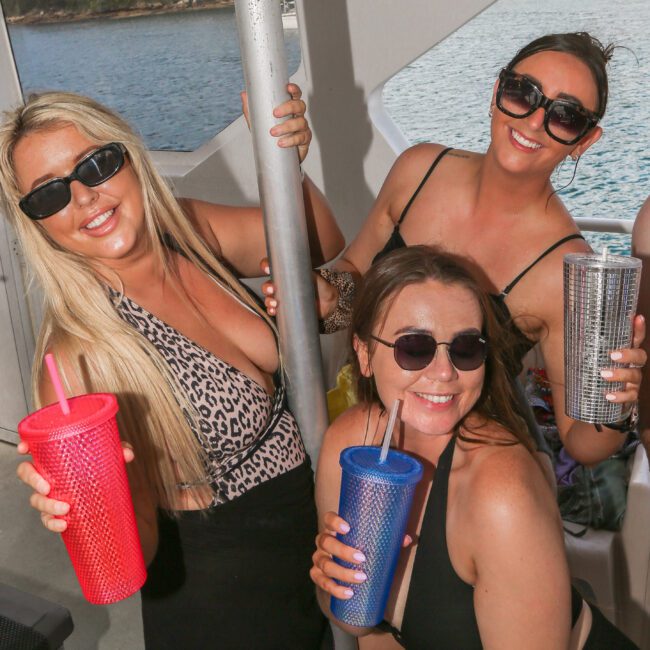 Three women in swimsuits and sunglasses smile while holding colorful tumblers with straws on a boat. They appear to be posing and enjoying a sunny day on the water, with a distant shoreline visible in the background.