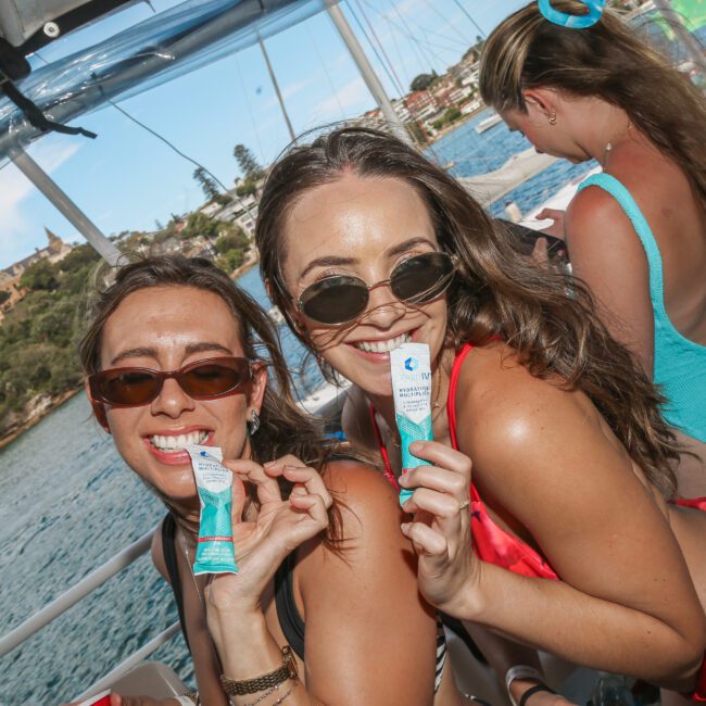 Two women on a boat smile at the camera, holding tubes of sunscreen. They wear sunglasses and swimwear. The background shows a scenic view of water and greenery. Other people in swimwear are visible on the boat.