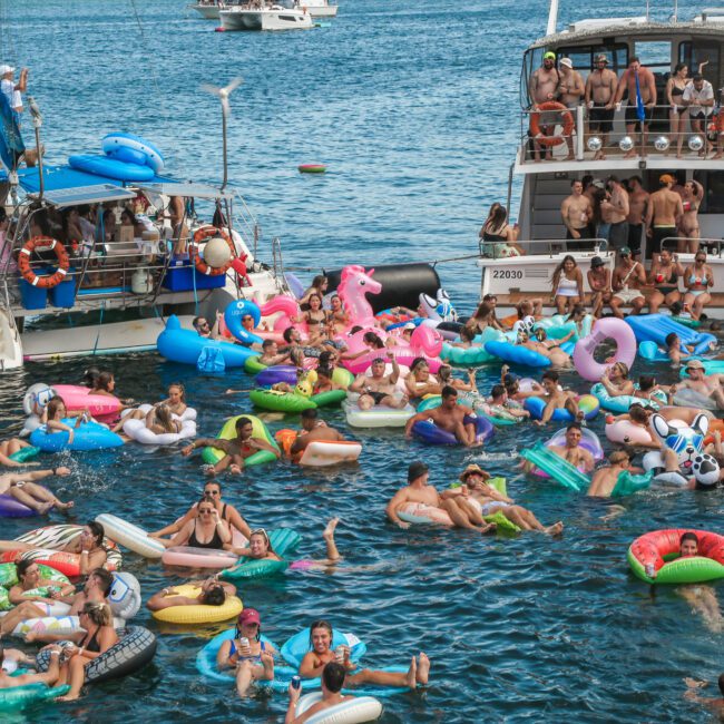 A crowded scene of people enjoying a sunny day in the water. Many are on colorful inflatable floats, including flamingos and unicorns. Two nearby boats are filled with people, and the vibrant blue sea adds to the lively atmosphere.