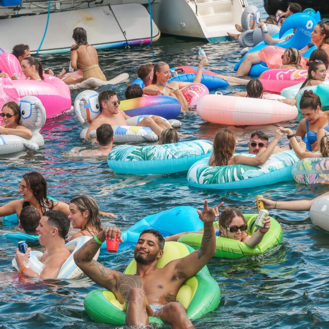 People are floating on colorful inflatables in the water near a boat. They are enjoying a sunny day, holding drinks and relaxing. The scene is lively and festive with a mix of palm and animal-shaped floats.