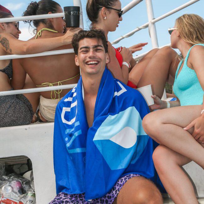 A group of people on a boat enjoying sunny weather. One man in the foreground is smiling, wrapped in a blue towel, and wearing purple leopard-print shorts. Others are sitting and chatting, dressed in swimwear. The sky is clear with some clouds.