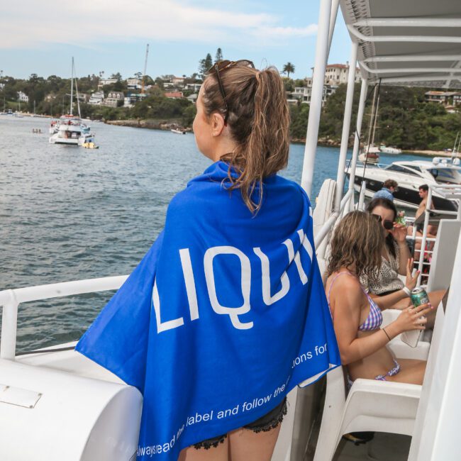 A person with wet hair stands on a boat, wrapped in a blue towel with white text. They're looking out at the water, where several boats are anchored. Another person sits nearby in a swimsuit. Houses and trees are visible on the distant shoreline.