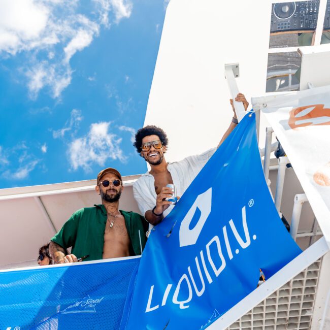 Two people on a boat, smiling and holding a large blue Liquid I.V. banner. One wears sunglasses and a hat, the other holds a drink. A clear sky with clouds is in the background.