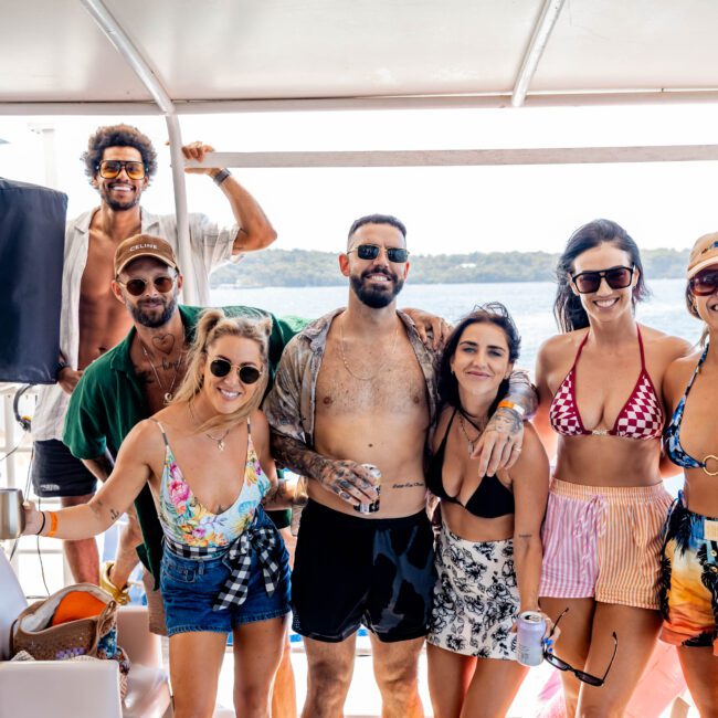 A group of seven friends in swimsuits pose together on a boat. They are smiling and standing close to each other. The ocean and distant land are visible in the background. Some wear sunglasses and hats, and a speaker is in view on the left.