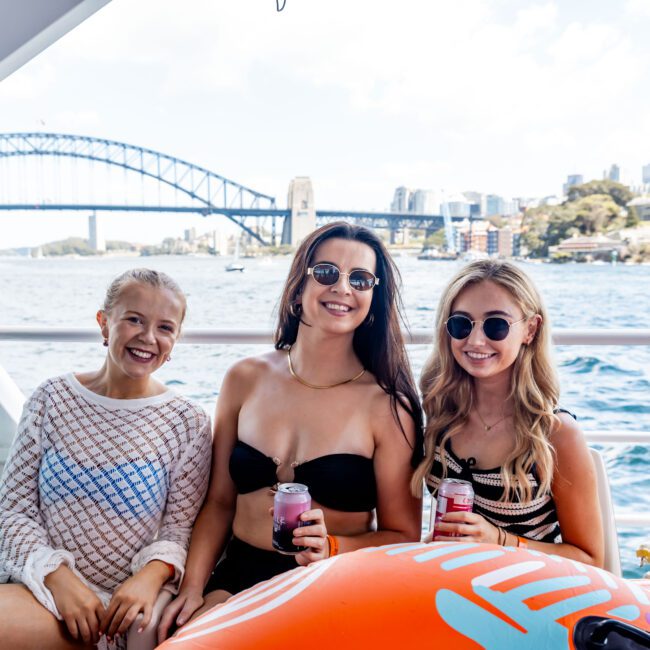 Three women seated on a boat with the Sydney Harbour Bridge visible in the background. They are smiling, wearing summer attire, and holding drinks. One is sitting on an inflatable orange item. The water and city skyline are visible.