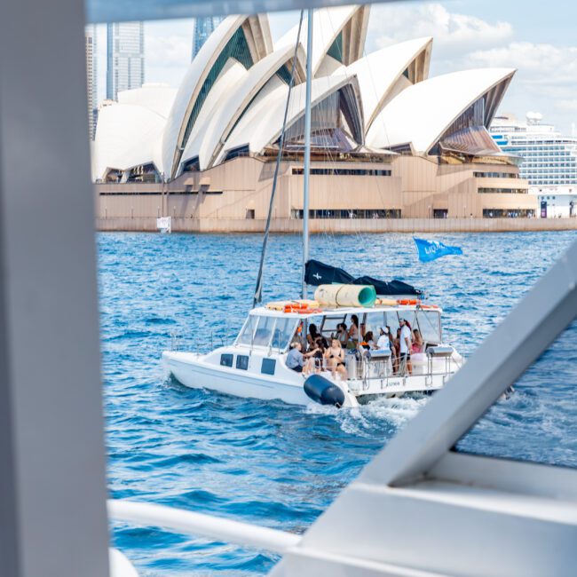 A group of people are enjoying a sunny day on a catamaran in a harbor, with the distinctive architectural design of the Sydney Opera House visible in the background. The scene is framed by parts of another boat.