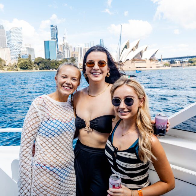 Three women smiling on a boat with the Sydney Opera House and Harbour Bridge in the background. They are dressed in casual attire, enjoying a sunny day on the water.