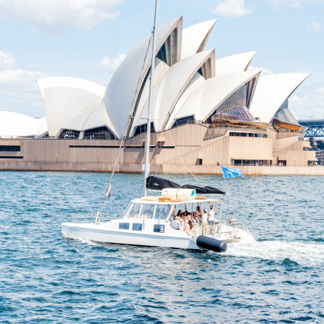 A white catamaran with passengers sails on water in front of the Sydney Opera House, with its iconic shell-like roof. The scene is set on a sunny day with a clear sky.