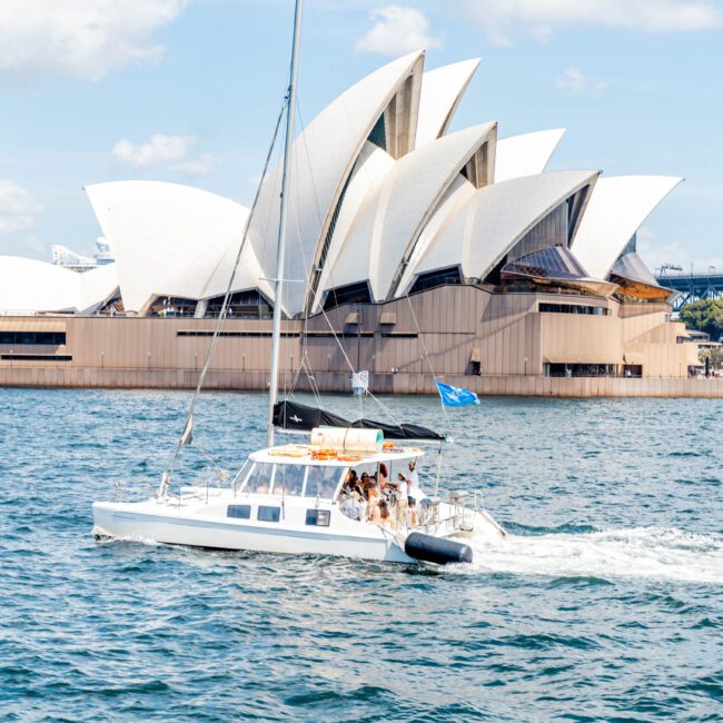 A small white sailboat with people on board glides through the water in front of the Sydney Opera House. The iconic building's white sails are prominent, and the Sydney Harbour Bridge is visible in the background under a blue sky.