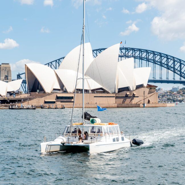 A sailboat navigates Sydney Harbour with the Sydney Opera House and Sydney Harbour Bridge in the background under a partly cloudy sky.