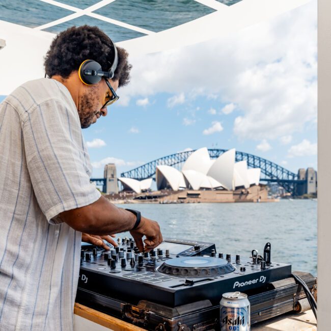 A DJ wearing headphones plays music on a mixer set up outdoors. In the background, the Sydney Opera House and Harbour Bridge are visible against a blue sky. A can of Asahi beer is on the table beside the equipment.