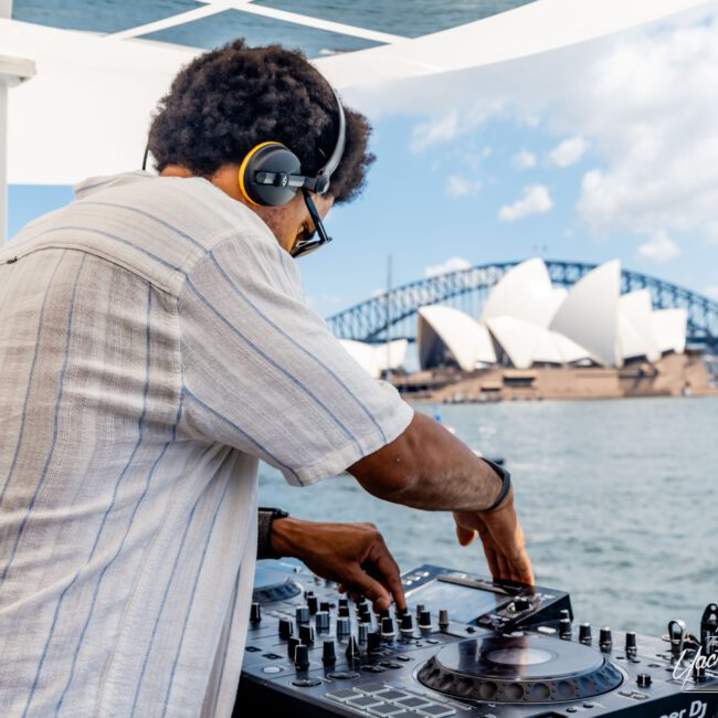 A DJ wearing a striped shirt and headphones is mixing music on a boat with a view of the Sydney Opera House and Sydney Harbour Bridge in the background. The scene is sunny and vibrant.