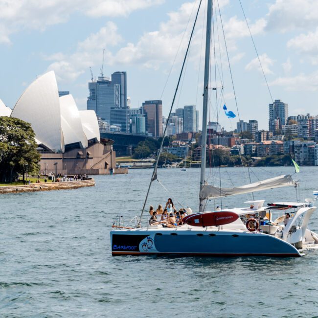 A sailboat with several people onboard cruises in front of the Sydney Opera House. The skyline of Sydney with high-rise buildings is visible in the background. The water is calm, and the sky is partly cloudy.