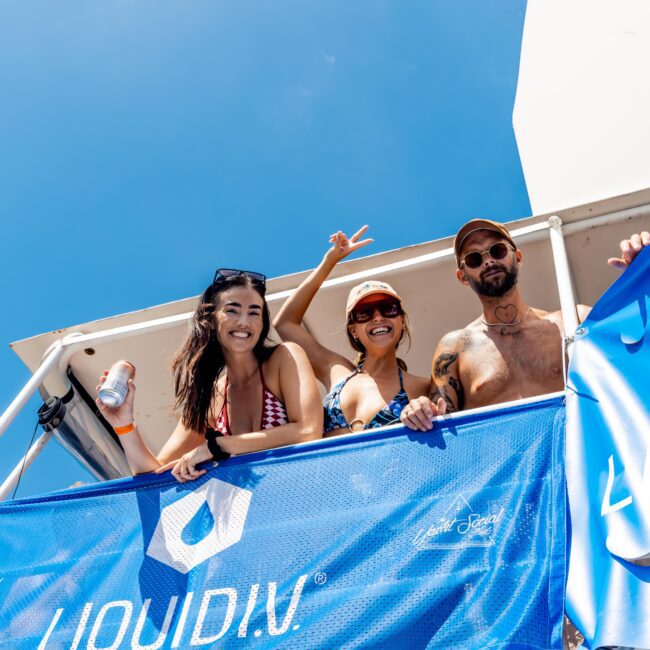 Three people stand on a boat deck, smiling and posing for the camera. The person in the middle shows a peace sign. A blue banner with "LIQUID I.V." is draped over the railing. The sky is clear and sunny.