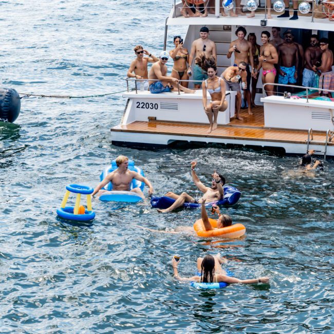 A group of people on a boat enjoy a sunny day. Several individuals are in the water on inflatable floats, waving and relaxing. The boat is large with people gathered along the railing, some holding blue flags. The scene is festive and lively.