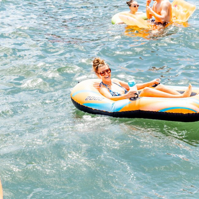 A woman in sunglasses and swimsuit lounges on an inflatable float in the water, holding a can. Other people relax on floats in the background on a sunny day.