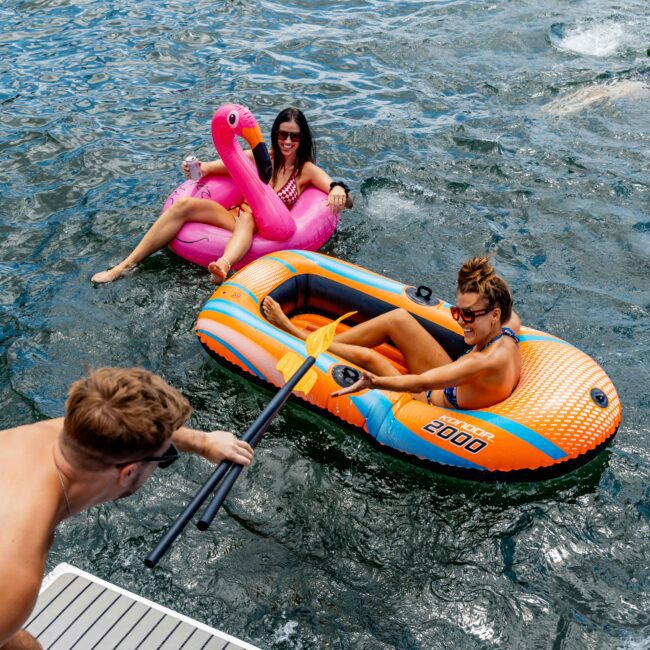 Two people are relaxing on inflatable floats in a body of water. One person is on an orange and blue raft, while the other is on a pink flamingo float. Another person is on a nearby dock reaching out with a paddle.