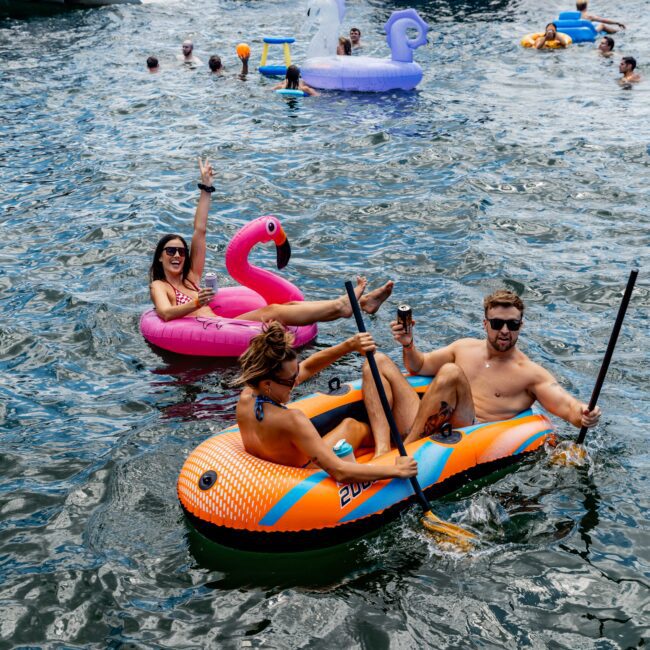 People enjoying a sunny day on a lake, lounging on inflatable rafts and floats. They hold drinks and paddle around near boats. Floats include a pink flamingo and swan. Other people swim and relax in the background under clear skies.