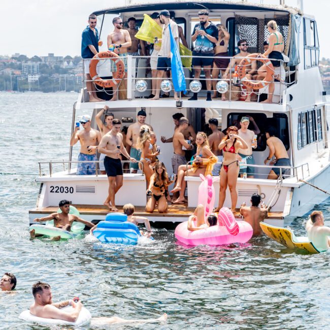 A group of people are enjoying a sunny day on a yacht and in the water. Some are on inflatable floats near the boat. The yacht is anchored in a body of water with a cityscape in the background. They are all in swimwear, and one person holds a flag.