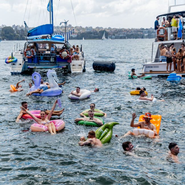 People float on inflatable toys in a lake near two boats. The scene is lively, with a variety of colorful inflatables shaped like animals and other objects. The background features a city skyline under a partly cloudy sky.