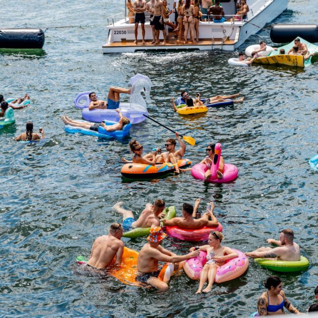 People enjoy a sunny day on a lake, floating on colorful inflatables like swans, rings, and loungers. Boats are nearby with more people onboard, adding to the lively and festive atmosphere.