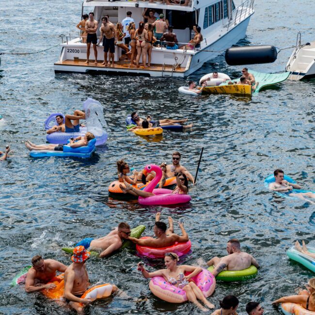 People enjoying a sunny day on a lake. A boat is in the background with people on board, while several others float nearby on inflatable rafts shaped like a swan, flamingo, and other colorful designs. The atmosphere is lively and festive.