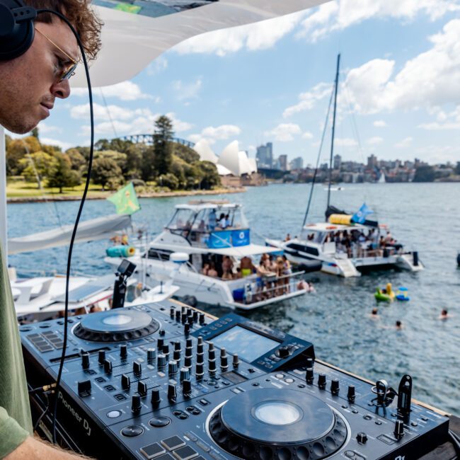 DJ wearing a green shirt and headphones plays music on a boat, with a backdrop of people enjoying themselves on nearby boats in the water. The sky is partly cloudy, and there are city buildings visible in the distance.