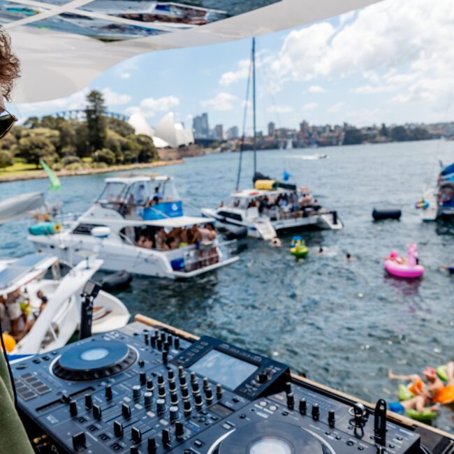 A DJ stands on a boat deck with audio equipment, overlooking a lively scene of people partying on nearby boats and inflatables in the water. The background features a city skyline and fluffy clouds.