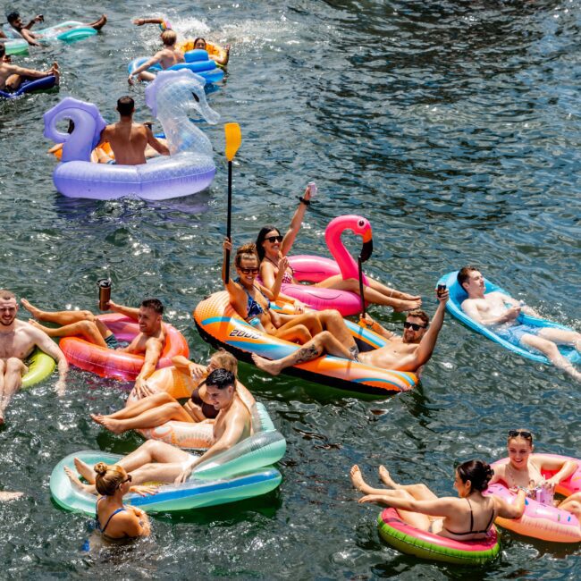 People relaxing on colorful inflatable floats in a sunny body of water. Floats include swans, flamingos, and lounge chairs. A yacht with people standing on it is in the background.