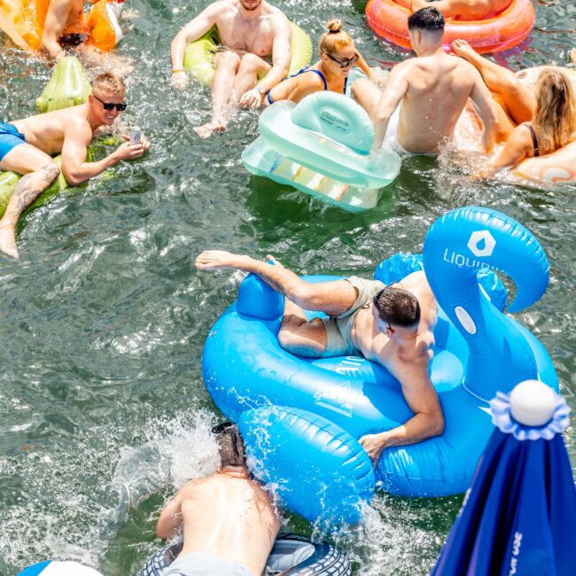 A group of people enjoying a sunny day on a lake, floating on colorful inflatable rafts, including a swan. Some are splashing water, and others are lounging. A large blue umbrella is visible in the foreground.