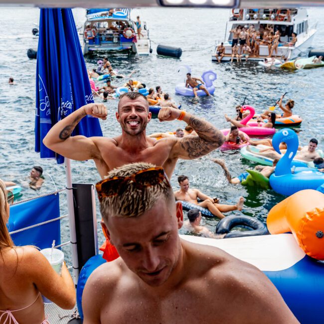 A man flexes his muscles and smiles on a boat surrounded by people swimming and lounging on colorful inflatables in the water. Boats filled with partygoers can be seen in the background on a sunny day.