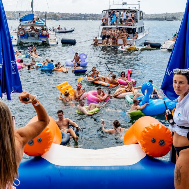 People enjoying a lively boat party. Many are in the water on colorful inflatables, while others dance on boats. Umbrellas are visible, and a slide leads into the water. The setting is a sunny day near a marina.