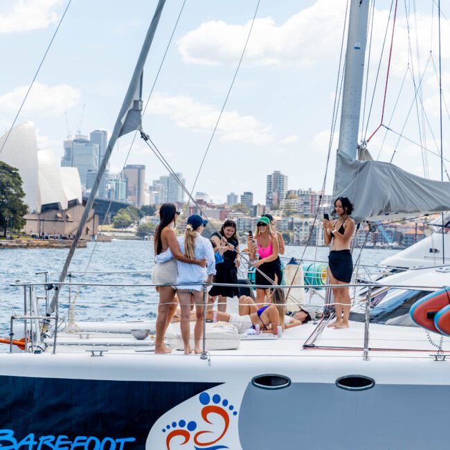 A group of people stands on a sailboat named "Barefoot" in a harbor with a city skyline and Opera House in the background. They appear to be socializing and enjoying the sunny day. The boat has colorful graphics on its side.